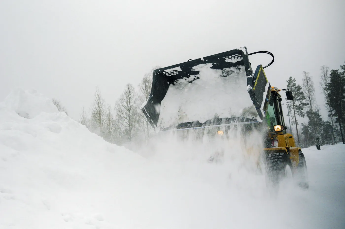 Gul lastartraktor med skopa flyttar snö vid en fastighet i djup vinter