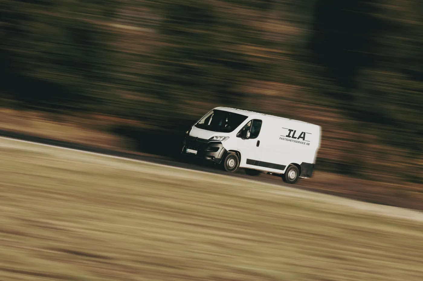 Vit servicebil med ILA Fastighetsservice-logotyp i fart på väg till uppdrag